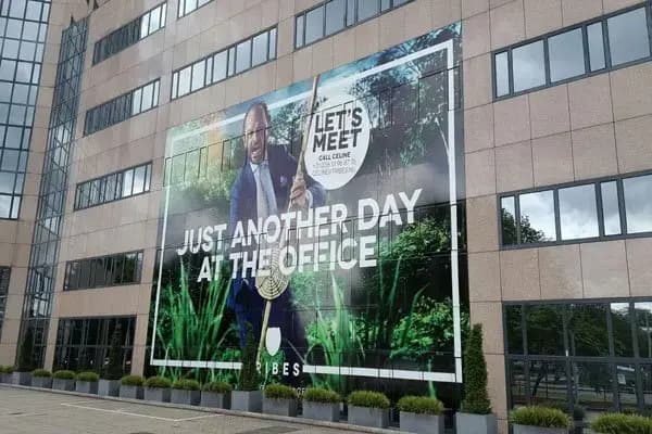 Kantoorgebouw met een gigantisch full-color geveldoek over meerdere verdiepingen, voorzien van een campagnebeeld en grote witte typografie “Just another day at the office”.
