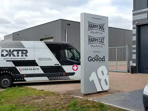 Vrijstaande reclametotem met merkbelettering voor diervoeding naast een zwart-wit bestickerde bedrijfsbus op een bedrijventerrein.