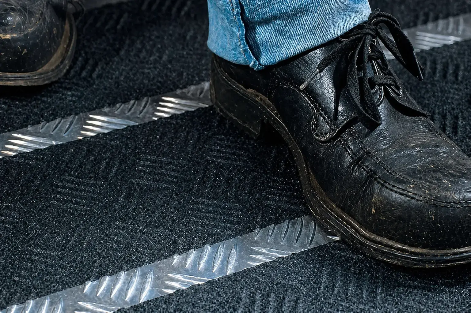Close-up van zwarte schoenen op een donkere vloermat met zilverkleurige antislipstrips, toegepast als veiligheidsmarkering en slippreventie op een looproute.