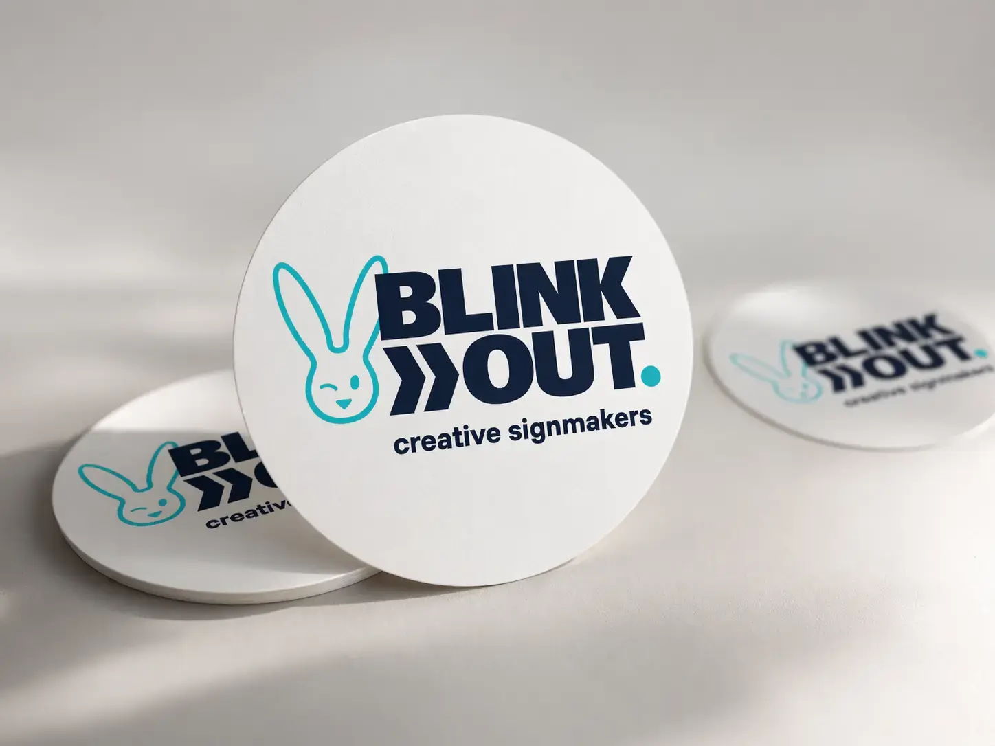 Witte ronde onderzetters met BLINK OUT creative signmakers-logo, gepresenteerd als promotioneel relatiegeschenk op een neutrale achtergrond.