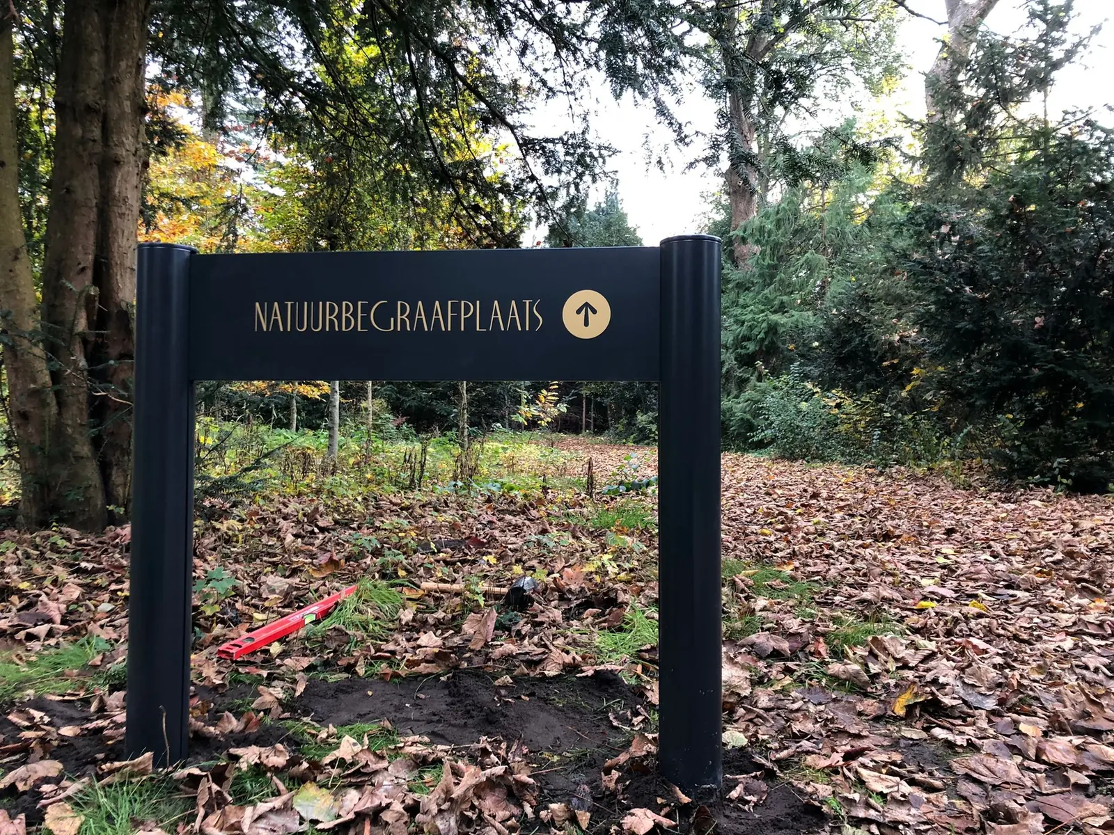 Dark directional sign with gold-coloured lettering for the natural burial ground and an arrow among trees in Hilversum.